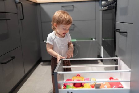 Téléchargez les photos : Portrait d'adorable enfant d'un an regardant à travers le tiroir de la cuisine, faible profondeur de champ - en image libre de droit