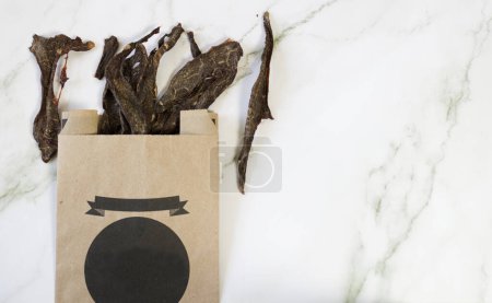 Foto de Dried meat in a recycled paper bag, healthy treats used for training or training a pet, especially dogs and cats, on a marble background - Imagen libre de derechos
