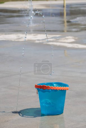 A child's blue bucket catches drips of water in this summertime splash pad and fountain