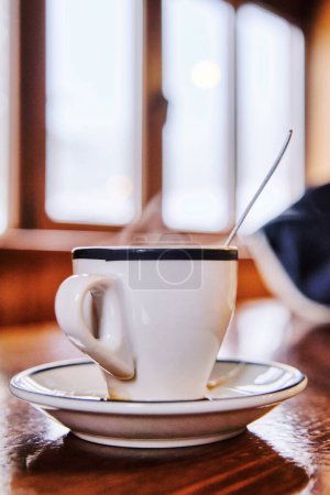 Foto de Taza de café expreso caliente con cuchara de café y plato en la mesa de madera con ventana en el fondo en el bar de montaña - Imagen libre de derechos