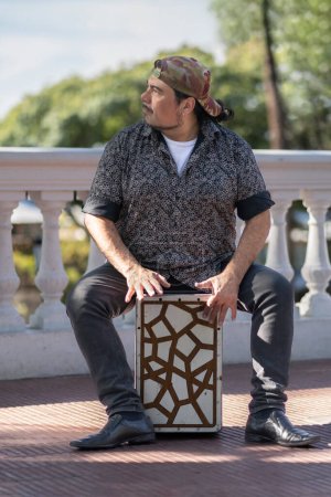 Foto de Hombre hispano le disparó a manos tocando percusión con una caja de flamenco sentada encima de ella en un parque - Imagen libre de derechos