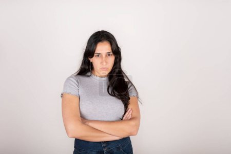 Portrait of a young latin woman dressed in a grey t-shirt with her arms crossed and an angry face, looking at the camera, on a white background