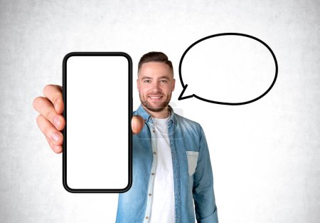 Téléchargez les photos : Portrait d'homme souriant montrant un grand smartphone simulant un écran d'espace de copie, bulle de parole vide sur fond de mur en béton gris. Concept de médias sociaux et d'applications mobiles - en image libre de droit