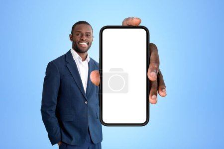 Photo for Smiling African American businessman wearing formal wear standing holding smartphone with mockup near empty blue wall in background. Concept of business person, mobile application, social media - Royalty Free Image