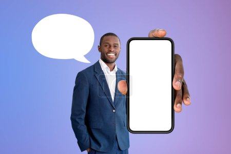 Photo for Smiling African American businessman wearing formal wear standing holding smartphone with mockup near purple wall with speech bubble in background. Presentation of mobile application, social media - Royalty Free Image