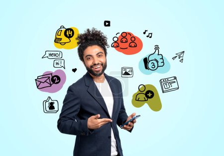 Téléchargez les photos : Sourire arabe homme d'affaires pointant vers smartphone, icônes doodle médias sociaux colorés sur fond bleu. Concept de communication et de marketing en ligne - en image libre de droit