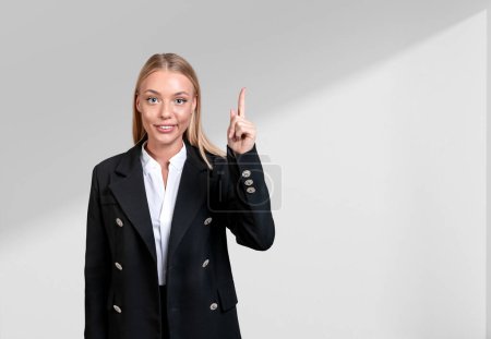 Foto de Mujer de negocios atractiva sonriente con ropa formal de pie sosteniendo su dedo hacia arriba cerca de la pared blanca vacía en el fondo. Concepto de persona de negocios ambiciosa, mujer inspirada, idea creativa - Imagen libre de derechos
