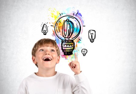 Photo for Impressed child boy with happy look and finger pointing up, bright light bulb and doodle on grey concrete wall. Concept of idea and plan - Royalty Free Image