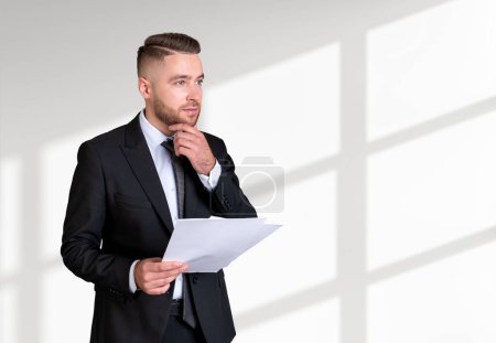 Photo for Dreaming handsome businessman wearing formal suit standing holding notes near empty white wall in background. Concept of model, pondering business person, considered man, lawyer, contract - Royalty Free Image