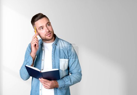 Photo for Dreaming handsome businessman wearing casual wear touch his forehead with pen and holding notebook near empty white wall in background. Concept of model, ambitious business person, student, learning - Royalty Free Image