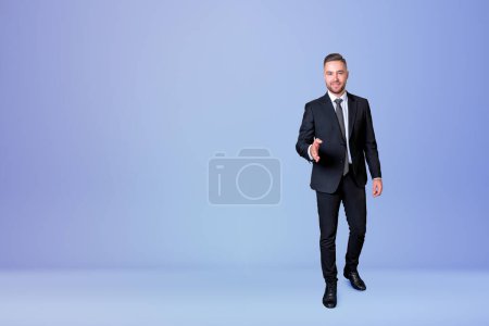 Foto de Smiling handsome businessman wearing formal suit standing lending hand for handshake near empty blue wall in background. Concept of ambitious business person, greeting gesture, successful inspired man - Imagen libre de derechos