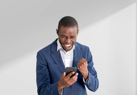 Foto de Feliz hombre de negocios afroamericano en traje formal sosteniendo teléfono inteligente y mostrando el puño cerca de la pared blanca vacía en el fondo. Concepto de persona de negocios ambicioso, gesto ganador, empresario exitoso - Imagen libre de derechos