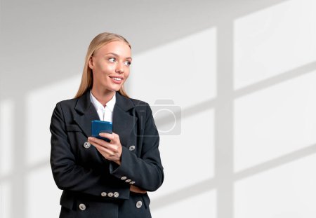 Photo for Inspired attractive businesswoman wearing formal wear standing in cross arm pose holding smartphone near empty white wall in background. Concept of ambitious business person, inspired woman - Royalty Free Image