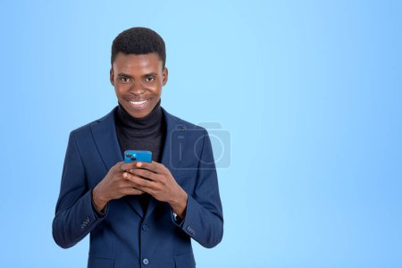 Photo for Smiling African American businessman wearing formal suit standing holding smartphone near empty blue wall in background. Concept of internet communication, mobile application, social media - Royalty Free Image