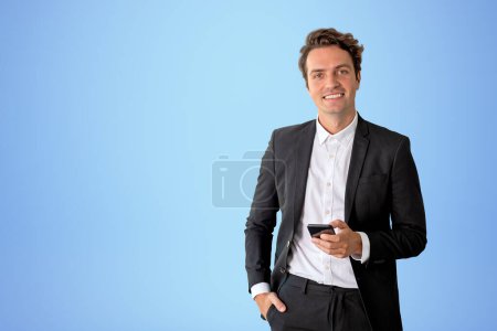 Foto de Smiling businessman working with phone, looking at the camera on copy space blue background. Concept of social media and online connection - Imagen libre de derechos