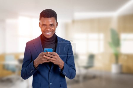 Photo for Smiling African American businessman wearing formal suit standing holding smartphone at office workplace in background. Concept of internet communication, mobile application, social media - Royalty Free Image