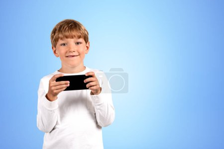 Photo for Smiling handsome boy in casual wear standing holding smartphone near empty blue wall in background. Concept of ambitious child, inspired kid, social media, mobile application, education, playing game - Royalty Free Image