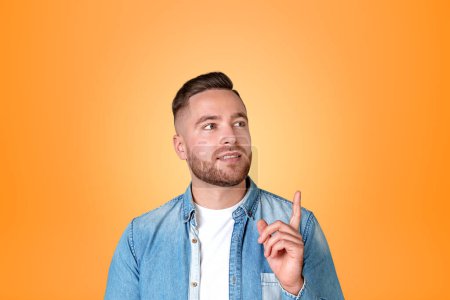 Photo for Pensive and smiling man in denim shirt, finger pointing up on copy space orange background. Concept of business offer and deal - Royalty Free Image