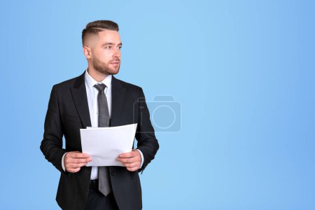 Photo for Dreaming handsome businessman wearing formal suit standing holding notes near empty blue wall in background. Concept of model, pondering business person, considered man, lawyer, contract - Royalty Free Image