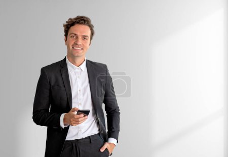 Téléchargez les photos : Happy businessman working with phone, hand in pocket looking at the camera. Copy space grey background. Concept of mobile app and online connection - en image libre de droit