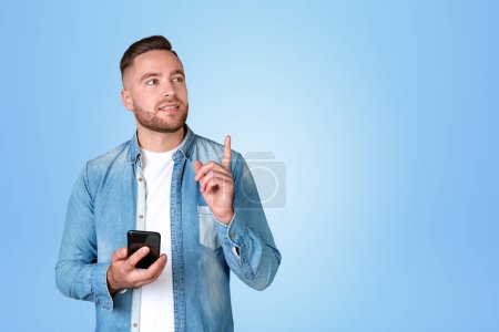 Téléchargez les photos : Inspired handsome businessman in casual wear standing holding smartphone near empty blue wall in background. Concept of ambitious business person, dreaming man, mobile application, social media - en image libre de droit