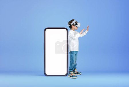 Photo for Child boy working in vr glasses, hands touching something in metaverse. Large mock up smartphone screen on blue background. Concept of mobile app and virtual reality - Royalty Free Image