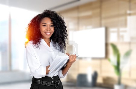 Téléchargez les photos : Smiling African American businesswoman wearing formal wear standing holding notebook taking notes at office workplace in background. Concept of student, successful business person, manager, learning - en image libre de droit