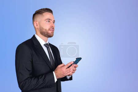 Téléchargez les photos : Inspired handsome businessman wearing formal wear standing holding smartphone near empty blue wall in background. Concept of pondering business person, dreaming man, social media, mobile application - en image libre de droit
