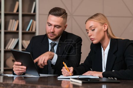 Photo for Pensive businessman and businesswoman working together, man finger touch tablet and woman taking notes in notebook. Concept of business education. - Royalty Free Image