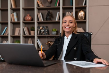 Photo for Happy businesswoman looking at the camera, sitting to desk with laptop and business document. Shelf with stylish decoration. Concept of business career and management - Royalty Free Image