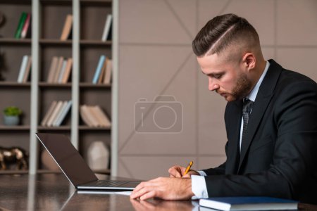 Téléchargez les photos : Pensive businessman working in office room with shelf and decoration, concentrated man profile with laptop on desk writing. Concept of distance work and education - en image libre de droit