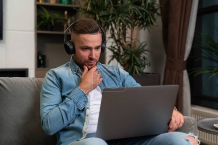 Téléchargez les photos : Pensive young businessman in headphones looking at laptop, hand on chin sitting on sofa at home. Concept of video conference and online education. - en image libre de droit