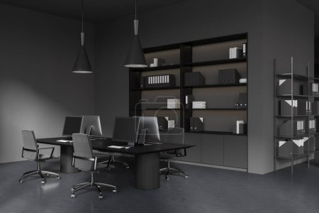Foto de Dark coworking interior with pc desktop on table and armchairs on grey concrete floor, side view. Business workplace with shelf and documents. 3D rendering - Imagen libre de derechos
