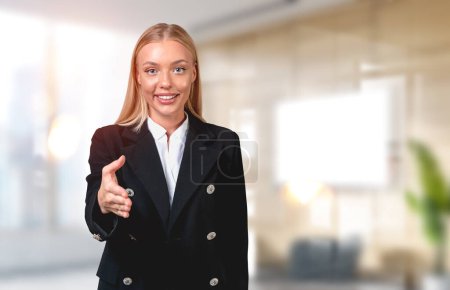 Téléchargez les photos : Smiling attractive businesswoman wearing formal wear standing stretching out hand for handshake at office workplace in background. Concept of ambitious business person, inspired woman - en image libre de droit