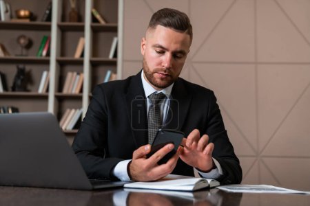 Foto de Serious handsome businessman wearing formal suit sitting typing on smartphone at office workplace with laptop in background. Concept of considered business person, pondering man, information, director - Imagen libre de derechos