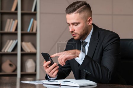 Foto de Serious handsome businessman wearing formal suit sitting typing on smartphone at office workplace in background. Concept of considered business person, pondering man, information, computer, director - Imagen libre de derechos