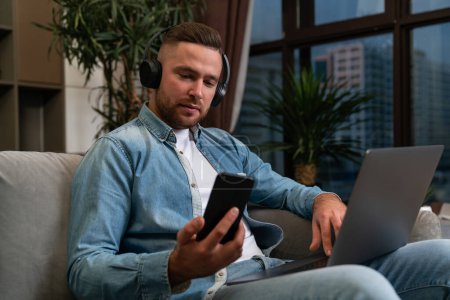 Téléchargez les photos : Handsome businessman in casual wear, headphones listening podcast on smartphone sitting on sofa with laptop on laps. Concept of self-education, online distant work, important conference call - en image libre de droit