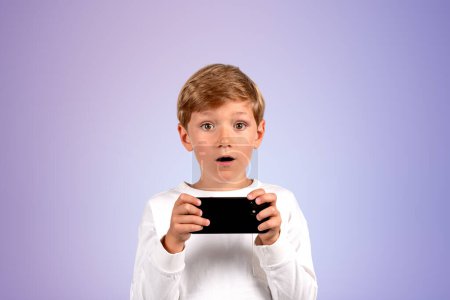 Téléchargez les photos : Shocked handsome boy in casual wear standing holding smartphone near empty purple wall in background. Concept of ambitious child, inspired kid, social media, mobile application, education, game play - en image libre de droit