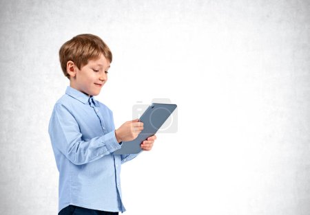 Photo for Smiling handsome boy in casual wear standing holding tablet device near empty concrete wall in background. Concept of ambitious inspired kid, social media, mobile application, education, playing game - Royalty Free Image