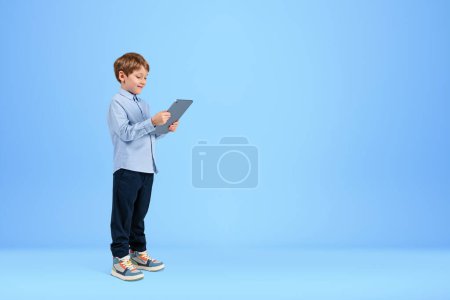 Foto de Smiling handsome boy in casual wear standing holding tablet device near empty blue wall in background. Concept of ambitious inspired kid, social media, mobile application, education, playing game - Imagen libre de derechos