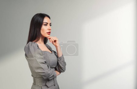 Photo for Young attractive businesswoman wearing formal wear is standing touching chin in cross arm pose near concrete wall with sun light in background. Concept of working process at workspace, brainstorming - Royalty Free Image