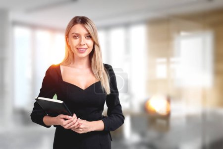Foto de Young attractive businesswoman wearing formal wear is standing holding notebook at office workplace with sun light in background. Concept of working process at workspace, work with documents - Imagen libre de derechos