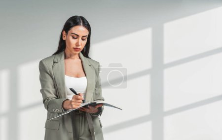 Téléchargez les photos : Businesswoman in beige suit, writing in business planner, pen in hand, grey background. Concept of business student and management - en image libre de droit