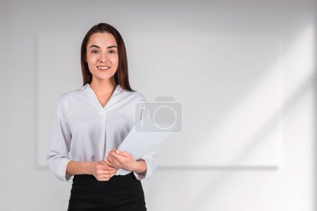 Téléchargez les photos : Smiling attractive businesswoman wearing formal wear standing holding pack of documents near empty white wall with mockup in background. Concept of ambitious business person, inspired lawyer - en image libre de droit