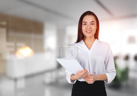 Téléchargez les photos : Smiling attractive businesswoman wearing formal wear standing holding pack of documents at office workplace in background. Concept of ambitious business person, inspired lawyer - en image libre de droit