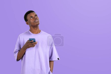 Foto de Pensive black man in t-shirt with phone in hand, thoughtful look up on empty copy space purple background. Concept of social media and communication - Imagen libre de derechos