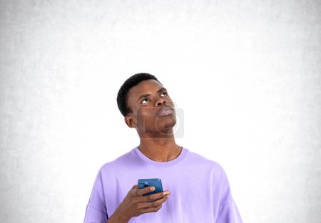 Foto de African man in purple t-shirt with phone in hand, thoughtful look up on empty grey concrete wall background. Concept of dream and idea - Imagen libre de derechos