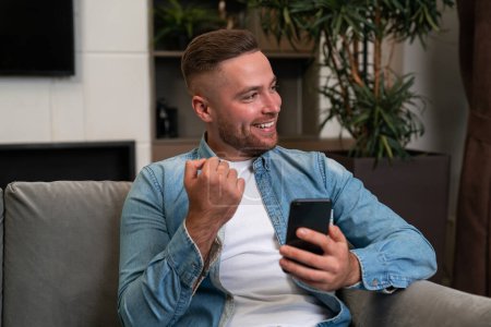 Photo for Glad handsome businessman wearing casual wear surfing net on smartphone sitting on sofa in background. Concept of smiling business person, pleased man, information, internet communication - Royalty Free Image
