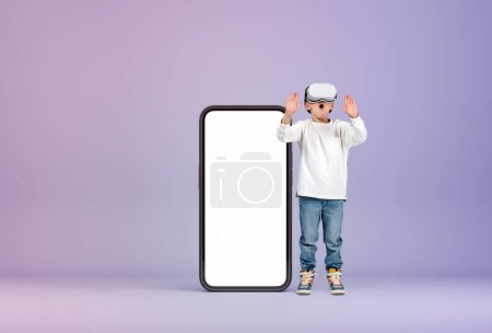 Photo for Excited child boy wearing vr glasses, touching something standing near large mockup empty phone screen. Concept of virtual world and metaverse - Royalty Free Image