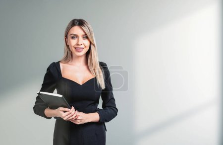 Téléchargez les photos : Jeune femme d'affaires attrayante portant des vêtements formels est debout tenant ordinateur portable près du mur de béton avec la lumière du soleil en arrière-plan. Concept de processus de travail sur l'espace de travail, travail avec des documents - en image libre de droit